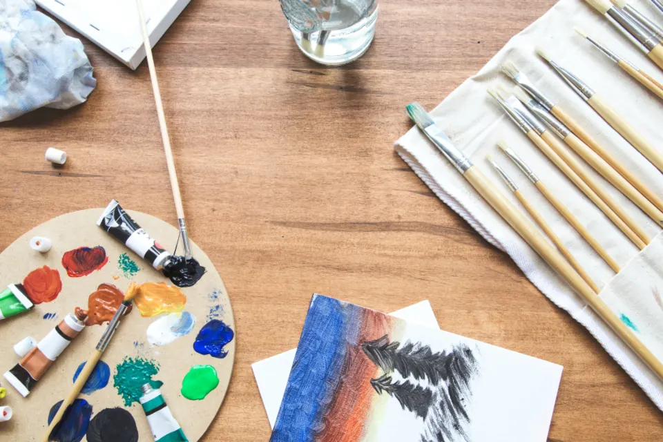 Paleta de pintor con pigmentos de colores y pinceles sobre una mesa de madera, junto a una ilustración empezada